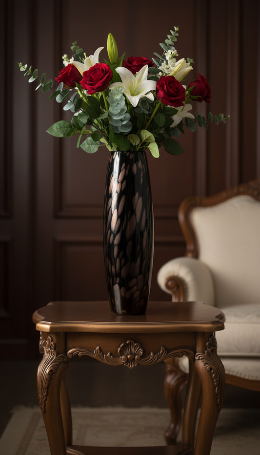 Espresso Blush Pebble Tall Column Vase lifestyle — 20.5 inch dark espresso hand-blown glass column with rose-blush pebble pattern and long-stemmed deep red roses, white lilies and eucalyptus on carved French baroque bronze side table in warm wood-panelled interior