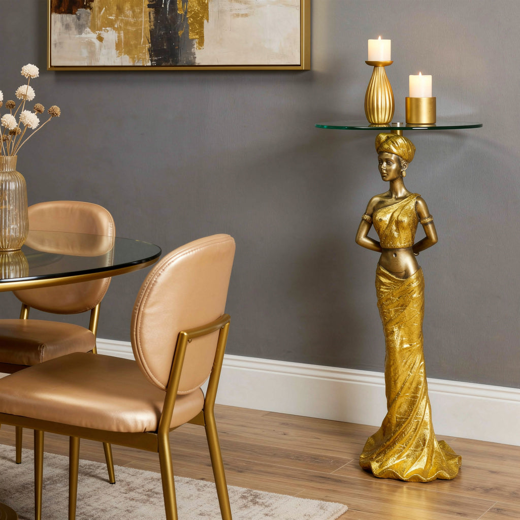 Gold African Queen Sculptural Glass Side Table lifestyle — 41 inch gold goddess pedestal with 12.5 inch round glass top and candles in golden and grey dining area corner with leather chairs and gold-framed abstract art
