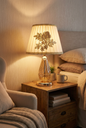 Rose Garden Crystal Table Lamp lifestyle — glowing warm gold light illuminating bedroom nightstand with ceramic mug, potted succulent and stacked books beside linen armchair and cozy bedding