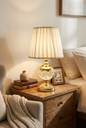 Gold Orb Crystal Table Lamp lifestyle — warm ivory honey glow on rustic oak bedroom nightstand beside stacked leather books and framed photograph with cream linen bedding and white wall background